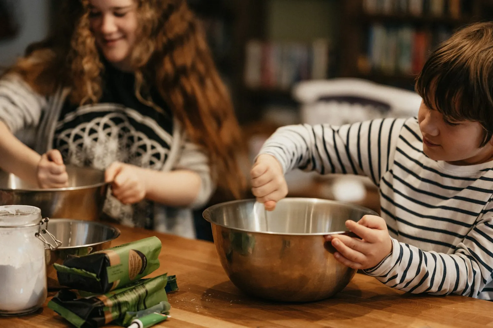 A boy and a young woman mixing batter in steel bowls at a wooden kitchen table at home
